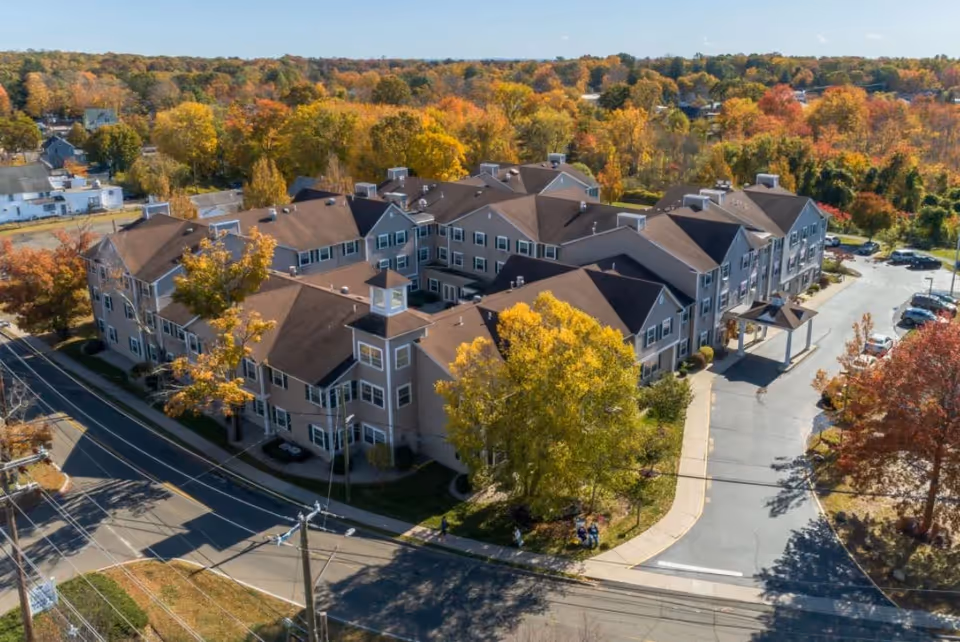 Aerial view of HarborChase of Madison senior living facility surrounded by trees with autumn foliage, showing a large multi-wing building with a brown roof and beige exterior, adjacent roads, parking areas, and a driveway entrance.