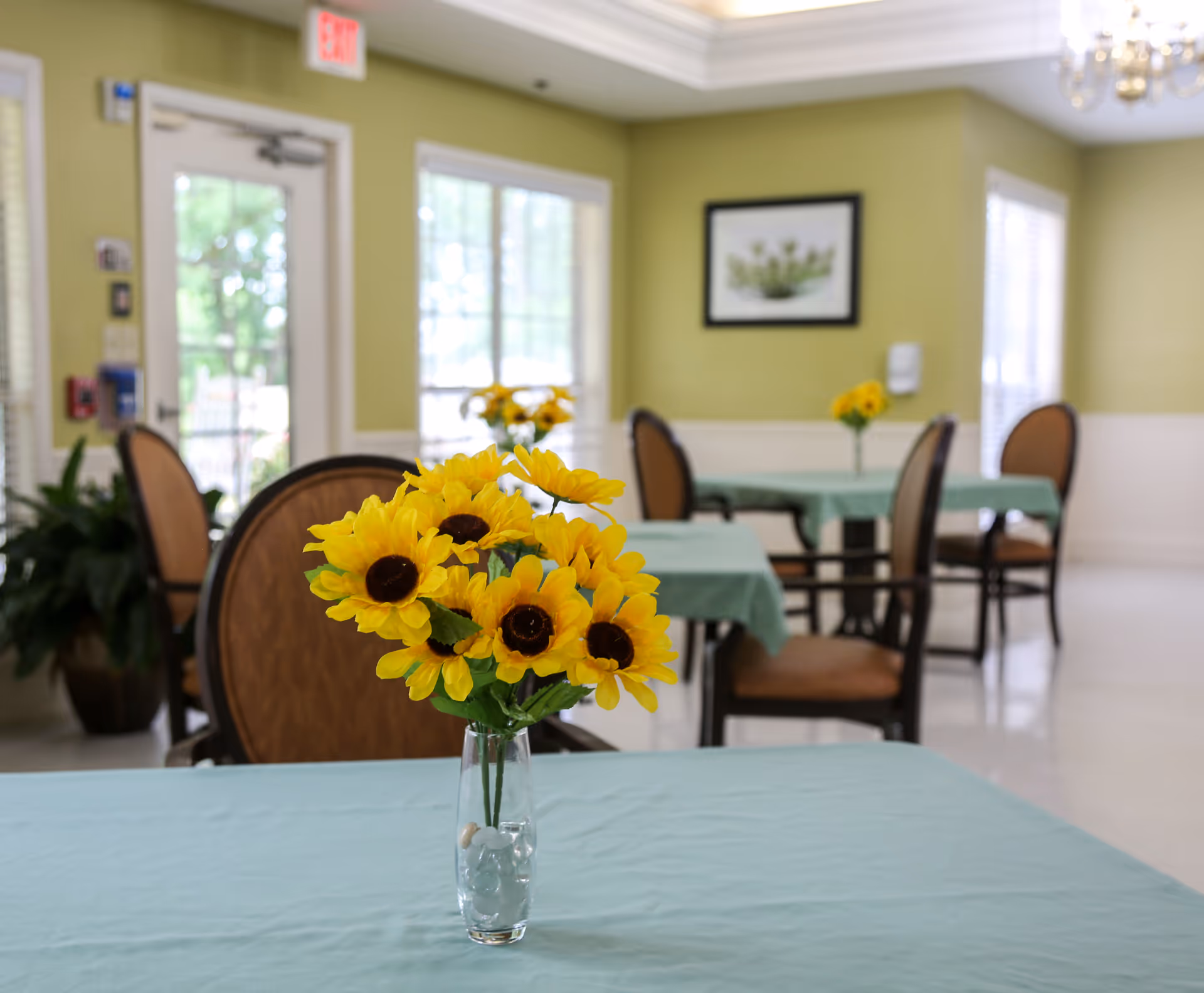 A bright dining room with tables covered in light blue tablecloths, each adorned with a vase of yellow sunflowers. The room has light green walls, large windows letting in natural light, and brown cushioned chairs around the tables.