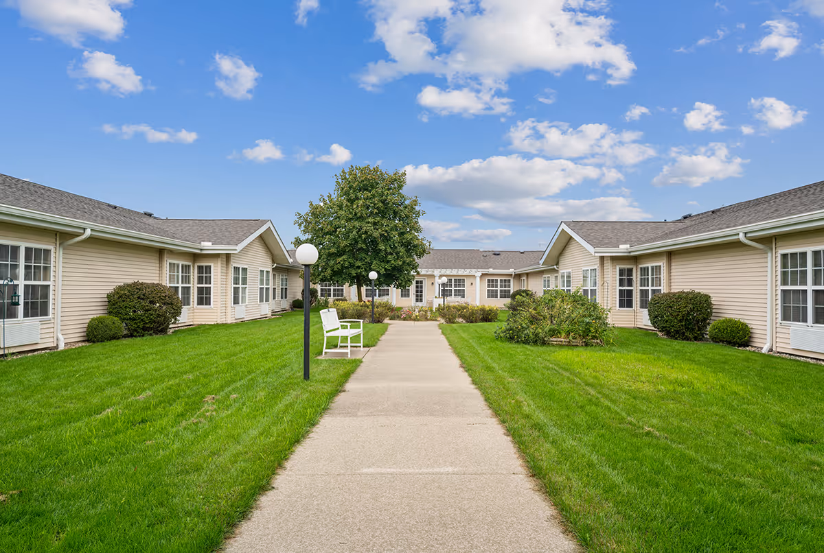 A paved walkway flanked by green lawns and bushes leads to a single-story beige building with multiple windows. There are white benches and lamp posts along the path under a partly cloudy blue sky.