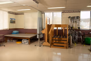 A rehabilitation room with a padded therapy table, stacked pillows, a set of wooden stairs with handrails for physical therapy, several walkers, and exercise equipment. The room has large windows with blinds and a beige and maroon color scheme.