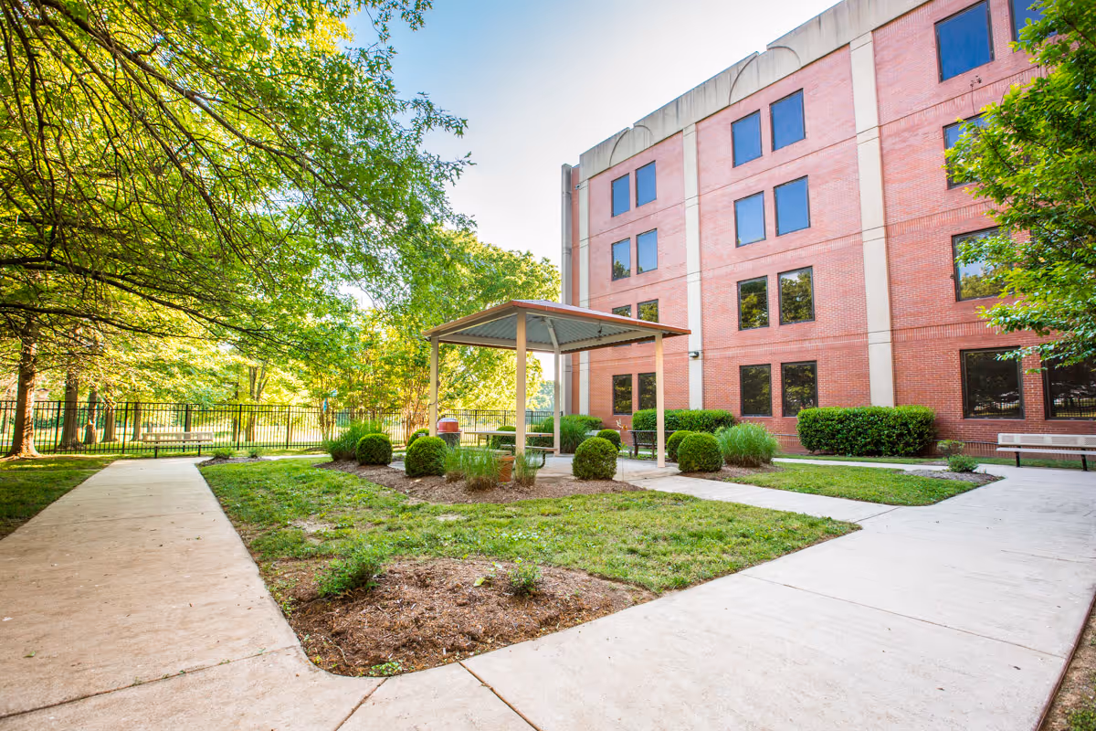 Outdoor courtyard area at Nashville Community Care & Rehabilitation at Bordeaux featuring a small covered pavilion with benches, surrounded by green bushes and trees, with a red brick building in the background and concrete walkways.