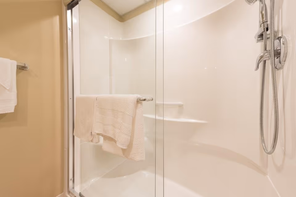 A clean, modern shower area with a glass sliding door. Inside the shower, there is a built-in seat and a handheld showerhead with a flexible hose. Two white towels hang on a towel bar attached to the glass door. The walls are light-colored, and the overall look is bright and minimalistic.