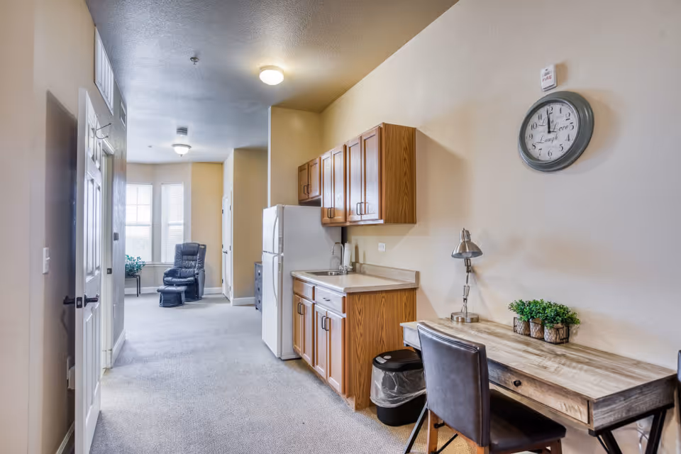 Interior view of a living space in an assisted living facility featuring a small kitchenette with wooden cabinets, a white refrigerator, and a sink. A wooden desk with a lamp, chair, and small potted plants is positioned against the wall beneath a round wall clock. In the background, there is a black recliner chair near windows letting in natural light.