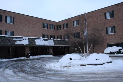 Brick senior living building with snow-covered driveway and entrance canopy, flags, and leafless trees in winter.