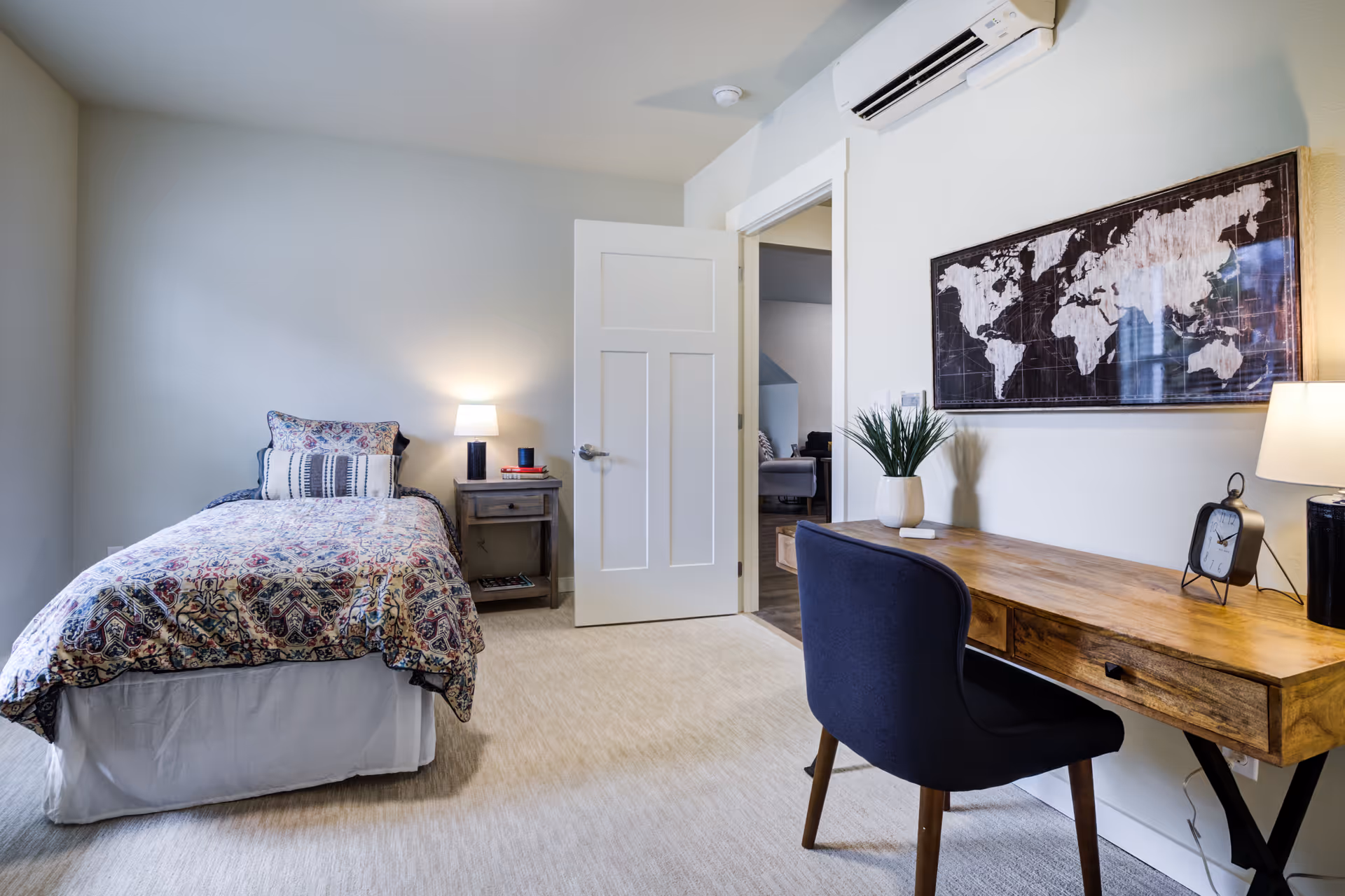 A cozy bedroom with a single bed covered in a patterned quilt and pillows, a wooden nightstand with a lamp and books, a wooden desk with a black chair, a potted plant, a clock, and a lamp. A world map artwork hangs on the wall above the desk. The door to the room is open, showing a glimpse of another room.