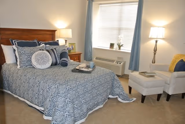 A cozy bedroom with a wooden headboard bed dressed in blue and white patterned bedding and multiple pillows. There is a wooden nightstand with a lamp and a framed photo beside the bed. A window with blue curtains and a white blind is behind an air conditioning unit. To the right, there is a beige armchair with a yellow pillow and a matching ottoman, both with a book on top. The room is softly lit by two lamps.