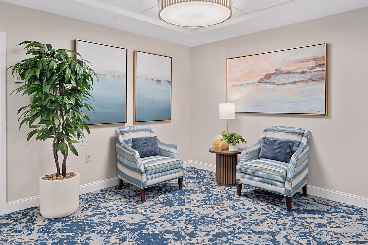 A cozy sitting area with two striped armchairs featuring blue cushions, a small round wooden side table with a lamp and a plant, a large potted green plant, and three abstract landscape paintings on the walls. The floor is covered with a blue and white patterned carpet.