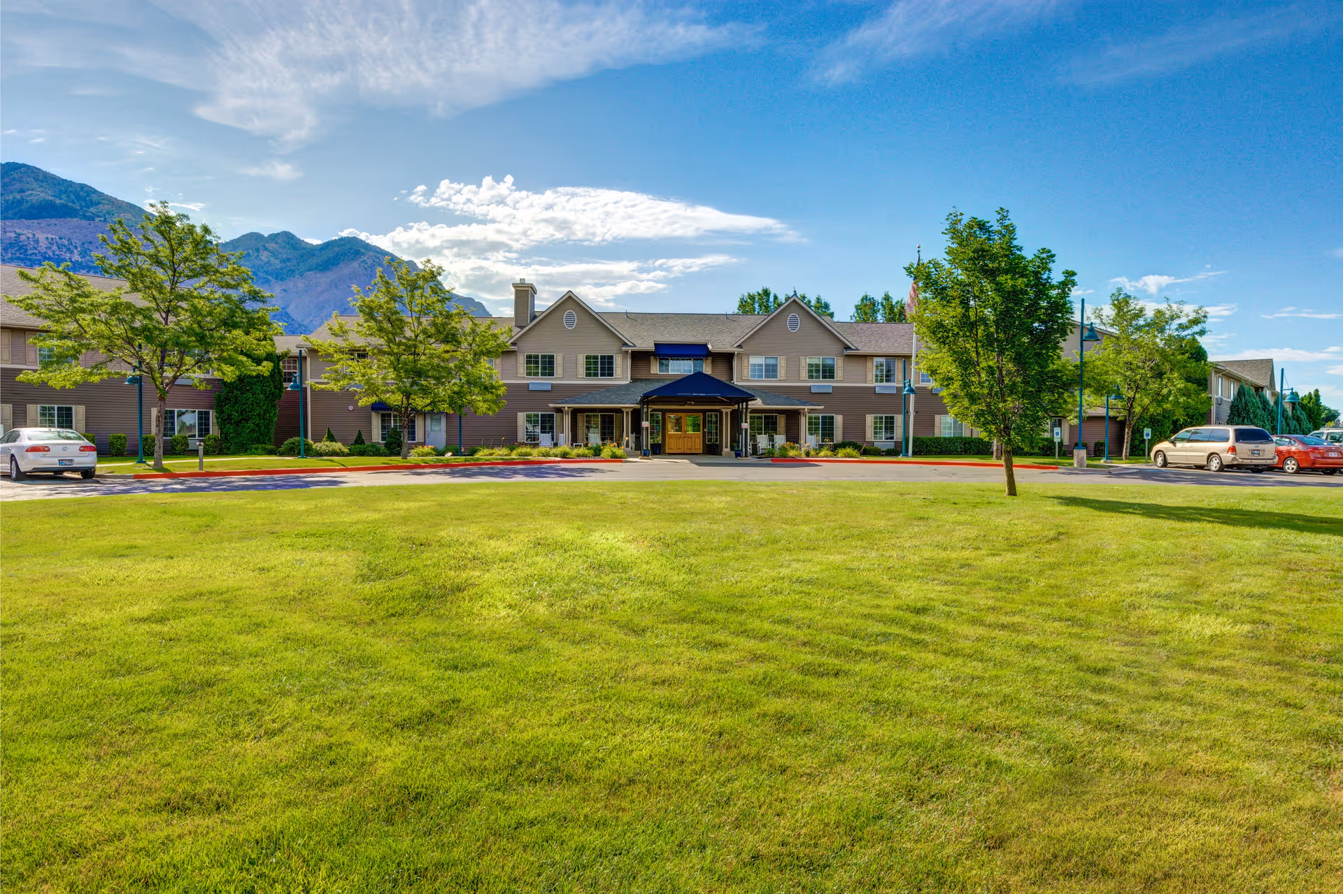Front exterior view of a two-story senior living facility building with a covered entrance, surrounded by green grass, trees, and parked cars under a blue sky with some clouds and mountains in the background.