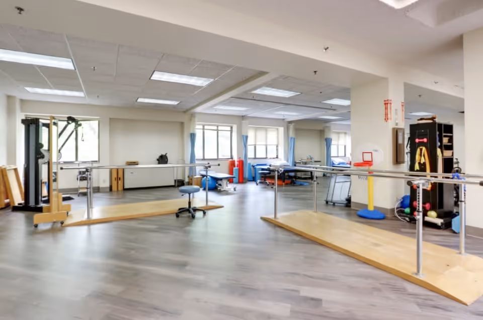 A spacious physical therapy or rehabilitation room with parallel bars for walking exercises, a stool, exercise equipment, therapy beds near windows with blue curtains, and various therapy tools and supplies organized around the room.