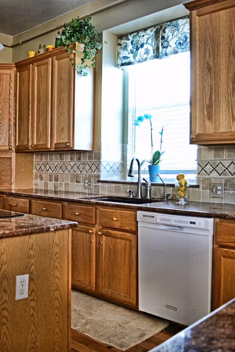Bright kitchen with oak cabinets, a window above a double sink, tiled backsplash, granite countertops and a dishwasher.