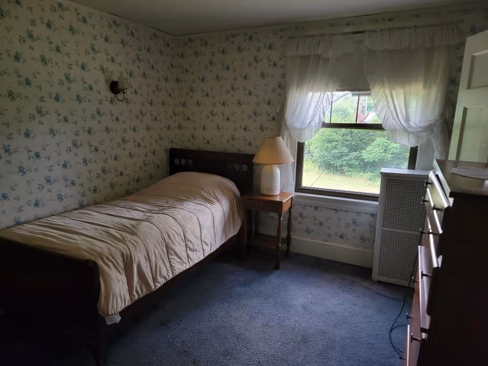 A small bedroom with floral wallpaper and blue carpet. The room contains a single bed with a beige comforter, a wooden nightstand with a lamp, a window with sheer white curtains, a radiator cover, and a wooden dresser.