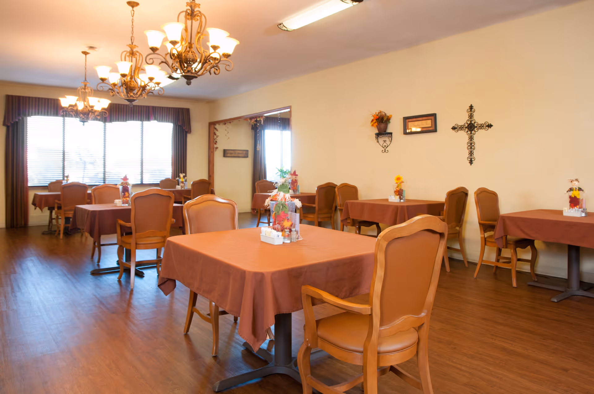 A dining room with several tables covered in brown tablecloths and surrounded by wooden chairs with padded seats and backs. The room has hardwood floors, large windows with blinds and valances, and decorative wall hangings including a cross and framed art. There are chandeliers hanging from the ceiling providing warm lighting.