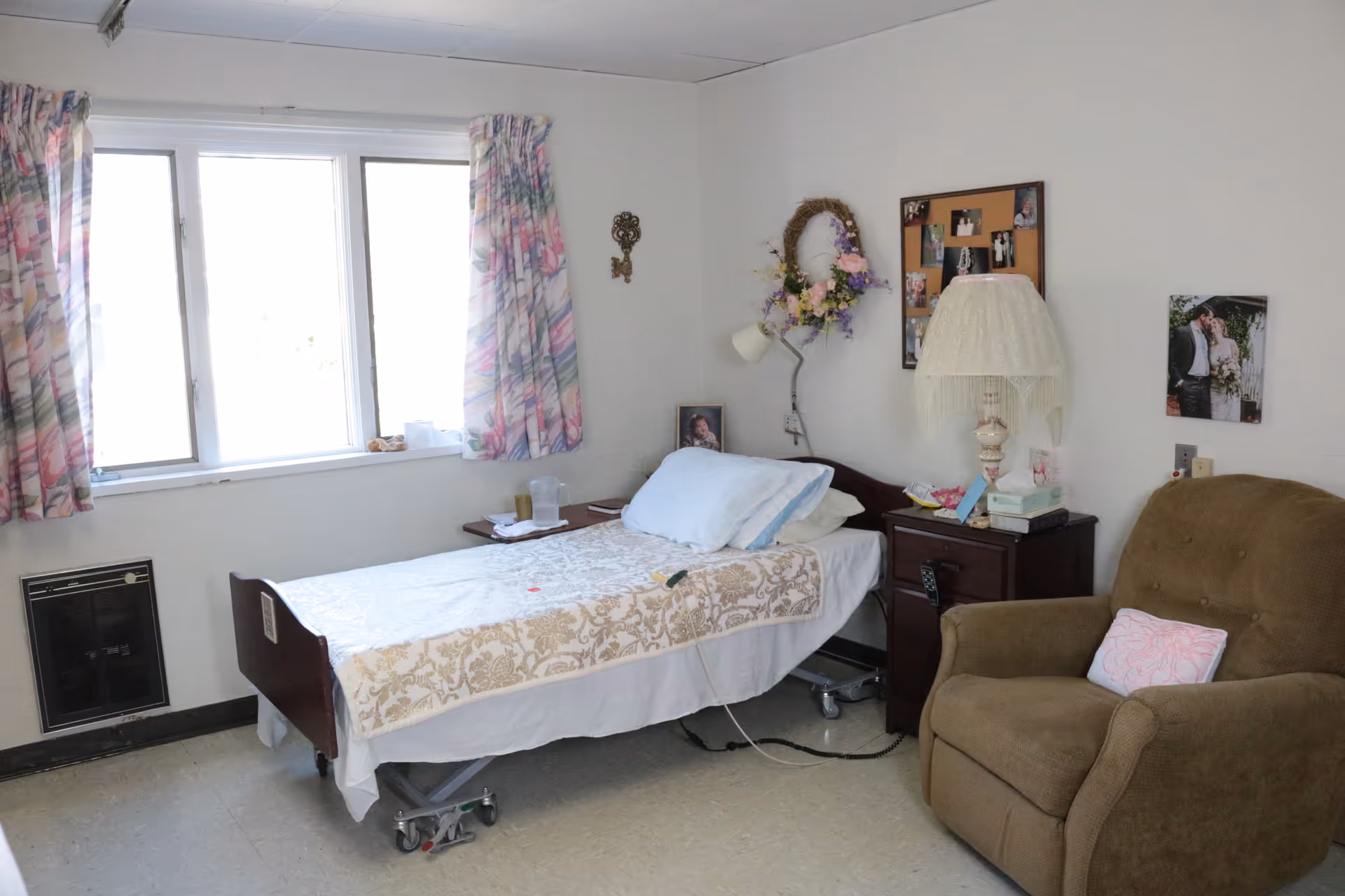 A small bedroom with a single hospital-style bed covered with a white and beige patterned blanket. Next to the bed is a wooden nightstand with a lamp, tissues, and some personal items. A brown recliner chair with a pink pillow is positioned nearby. The walls are decorated with a floral wreath, a bulletin board with photos, and a wedding photo. A window with floral curtains lets in natural light.