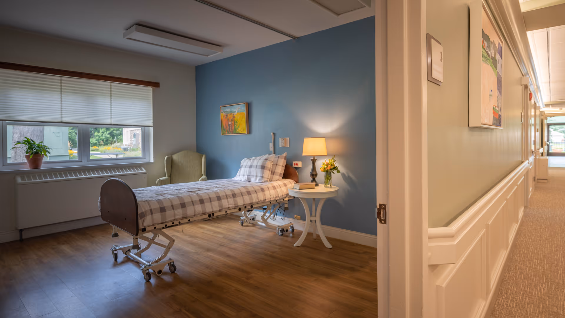 A bright senior living bedroom with a single adjustable bed, a bedside table and lamp, an armchair by the window, and a hallway visible outside the open door.