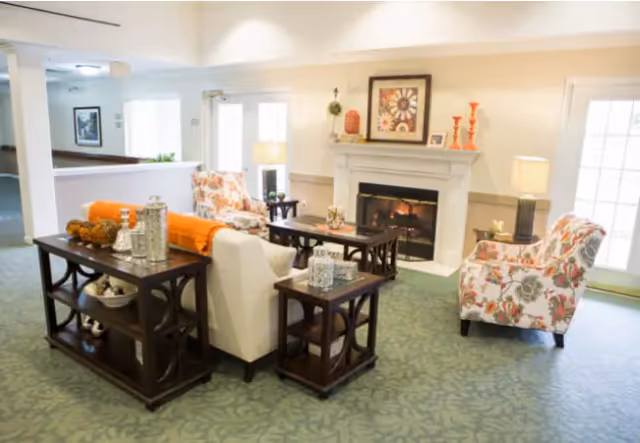 A cozy senior living room with a white sofa, two floral armchairs, a fireplace with decorative items on the mantel, and several dark wood tables with decorative pieces. The room has a green patterned carpet and is well-lit with natural light coming through glass doors.