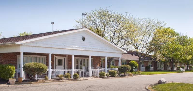 Exterior view of Belmont Terrace Nursing and Rehabilitation Center showing a single-story brick building with a white porch, columns, and a paved driveway. Trees and shrubs surround the building under a clear sky.