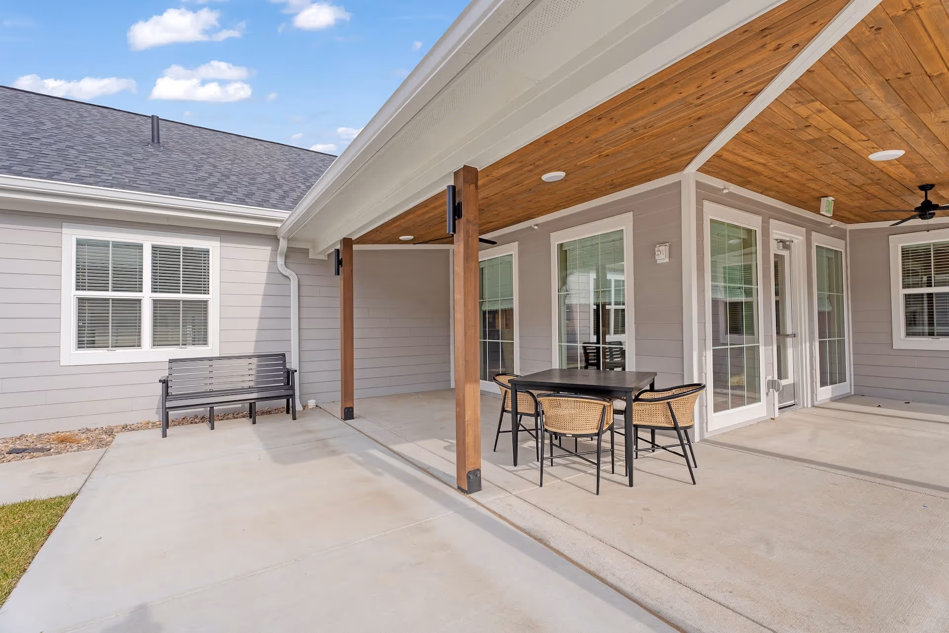 Outdoor covered patio area at Cedarhurst Senior Living of Salina with a black metal table and four wicker chairs, wooden ceiling with ceiling fans, a black bench against the building wall, and large windows and glass doors leading inside.