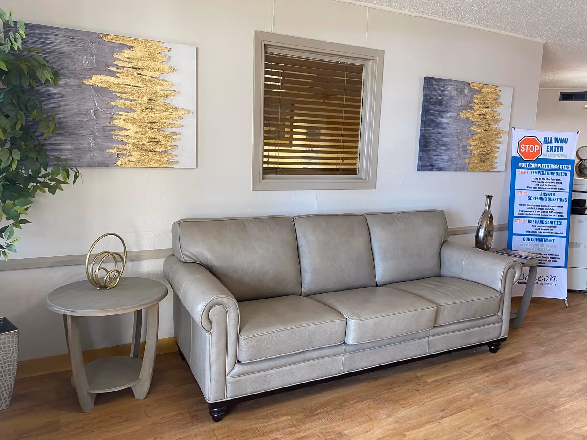 A beige leather sofa with three cushions is placed against a wall with a window featuring wooden blinds. On either side of the sofa are small round wooden tables, one with a decorative gold sculpture and the other with a tall silver vase. Two abstract paintings with gold and gray tones hang on the wall above the sofa. A green leafy plant is visible on the left side. On the right side, there is a standing sign with COVID-19 safety instructions. The floor is wooden.