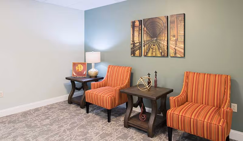 A cozy seating area with two orange and red striped armchairs separated by a dark wooden side table with decorative items. Another dark wooden side table with a lamp and a framed picture is next to one of the chairs. The wall behind is painted green and features a three-panel artwork depicting a library interior.
