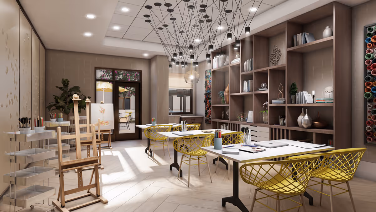 A bright and modern art studio room with wooden easels, white tables, and yellow wireframe chairs. The room features a large wooden shelving unit filled with books, decorative items, and art supplies. There are multiple pendant lights hanging from the ceiling, and a door leading to an outdoor patio area with yellow umbrellas is visible in the background.