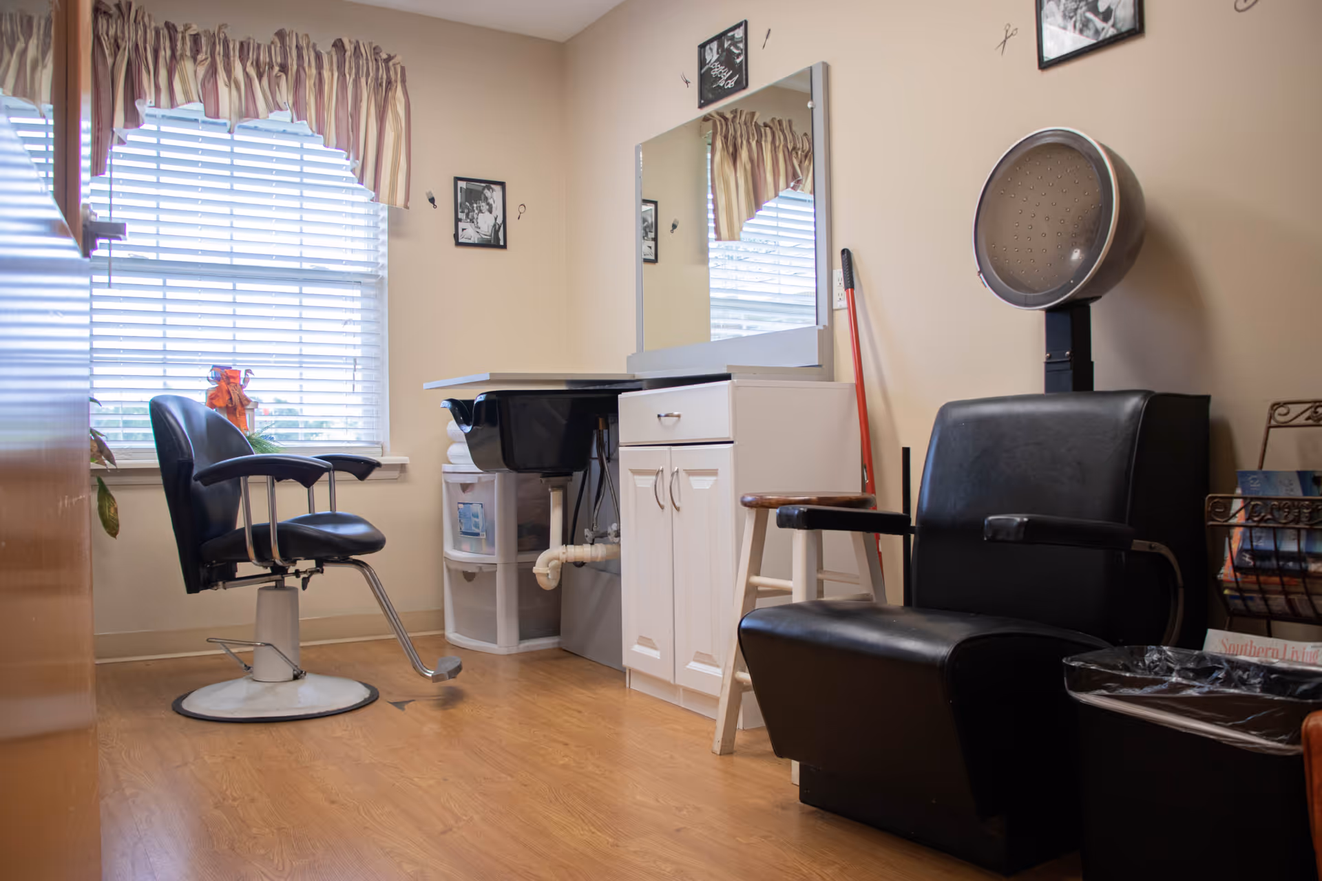 Interior of a small salon room with a black salon chair in front of a window with blinds and a striped valance. There is a white cabinet with a mirror above it, a black hair dryer chair, a small stool, and a magazine rack with magazines on the right side.