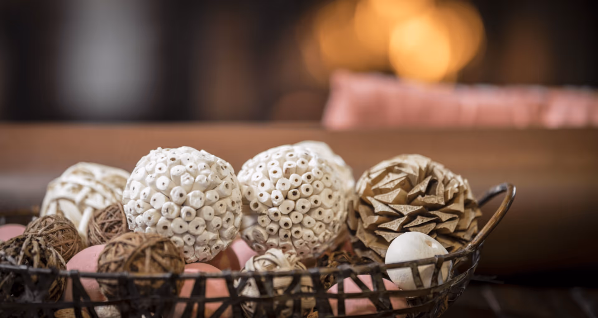 A decorative wire bowl filled with assorted textured decorative spheres sits on a table with a blurred interior background.
