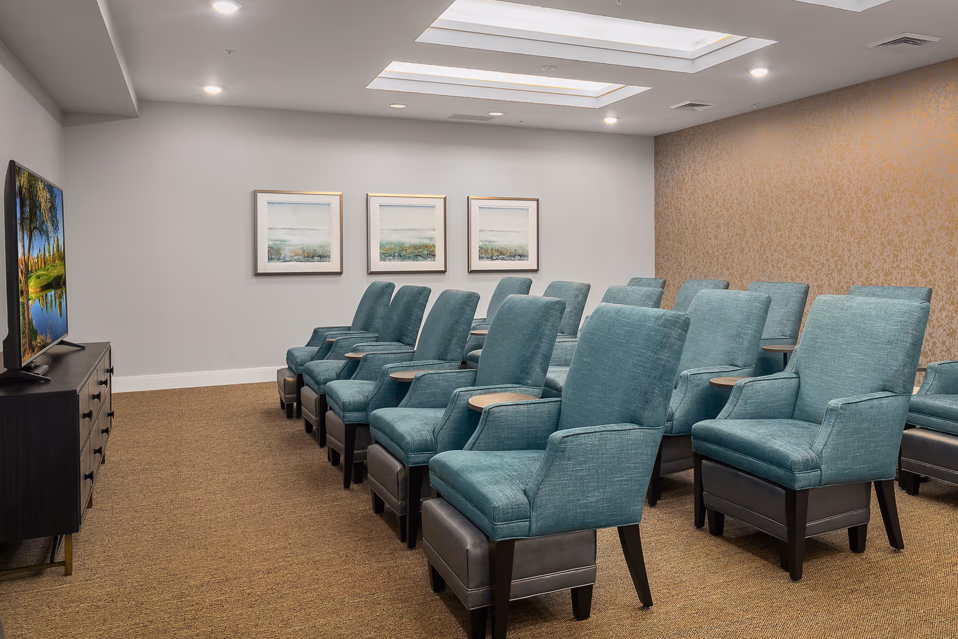 A small theater or media room with two rows of teal upholstered chairs, each with a small attached wooden table. A flat-screen TV is mounted on a dark wooden cabinet on the left side of the room. The walls are light gray with three framed landscape paintings and one wall covered in a textured golden wallpaper. The ceiling has recessed lighting and a skylight.