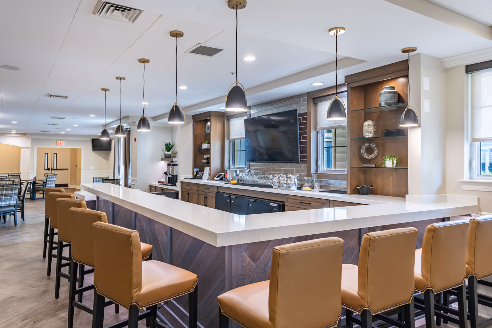 A modern dining area in a senior living facility featuring a long white countertop bar with tan leather bar stools. Pendant lights hang above the bar, and there is a large flat-screen TV mounted on the wall behind the bar. Shelves with decorative items and windows with blinds are also visible. In the background, there are additional dining tables and chairs.