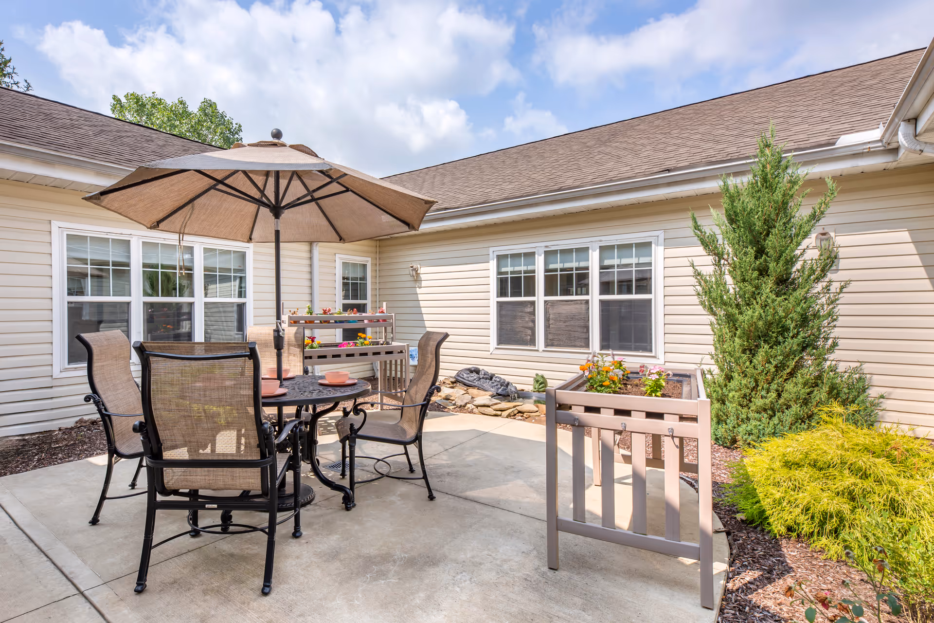 Outdoor patio area with a round table, four chairs, and a large umbrella. There are two raised garden beds with flowers and plants, surrounded by beige siding walls and windows of the building. The sky is partly cloudy.