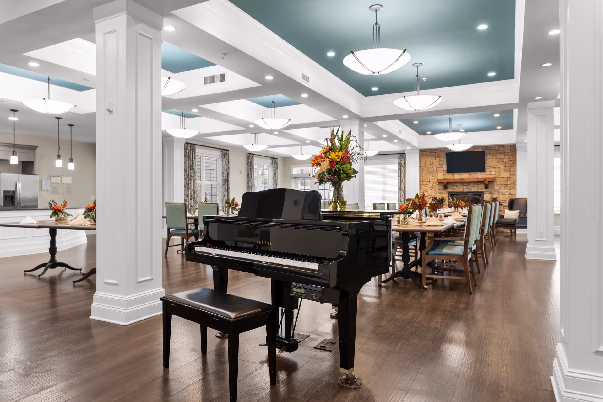 Spacious and elegant common area in a senior living facility featuring a black grand piano with a matching bench in the foreground, a large dining table set with napkins and floral centerpieces, a stone fireplace with a mounted TV, and a kitchen area with stainless steel appliances in the background. The room has wooden flooring, white columns, and a ceiling with recessed lighting and modern pendant lights.