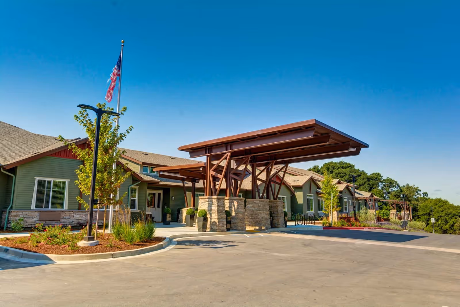Exterior view of The Vineyard at Fountaingrove Memory Care facility showing a single-story building with green siding and stone accents, a large covered entrance with wooden beams, landscaped plants, an American flag on a flagpole, and a clear blue sky.