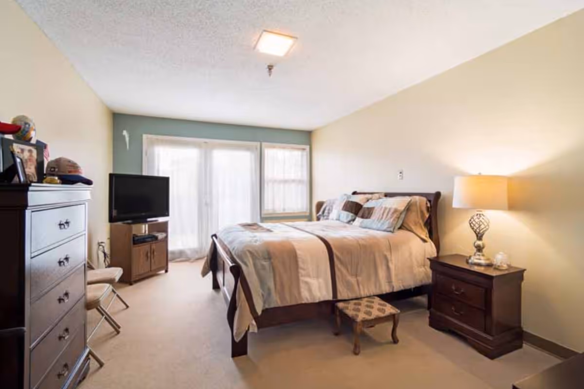 A well-lit bedroom with a large bed covered in beige and brown bedding, a wooden nightstand with a lamp, a small footstool at the bed's foot, a wooden dresser with personal items on top, a TV on a stand, and a window with sheer curtains letting in natural light.