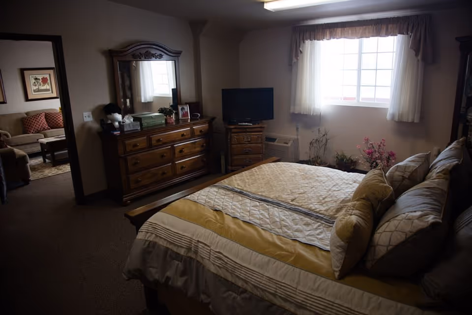 A cozy furnished bedroom with a made bed, multiple pillows, a wooden dresser with mirror, a small TV, and a window.