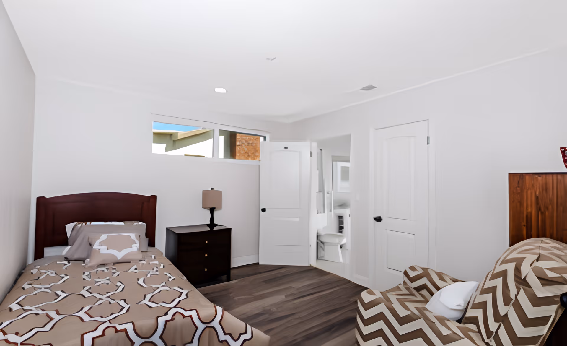 A small bedroom with a single bed covered in a beige and white patterned bedspread, a dark wooden nightstand with a lamp, a chevron-patterned armchair, and two white doors, one of which is open showing a glimpse of a bathroom.