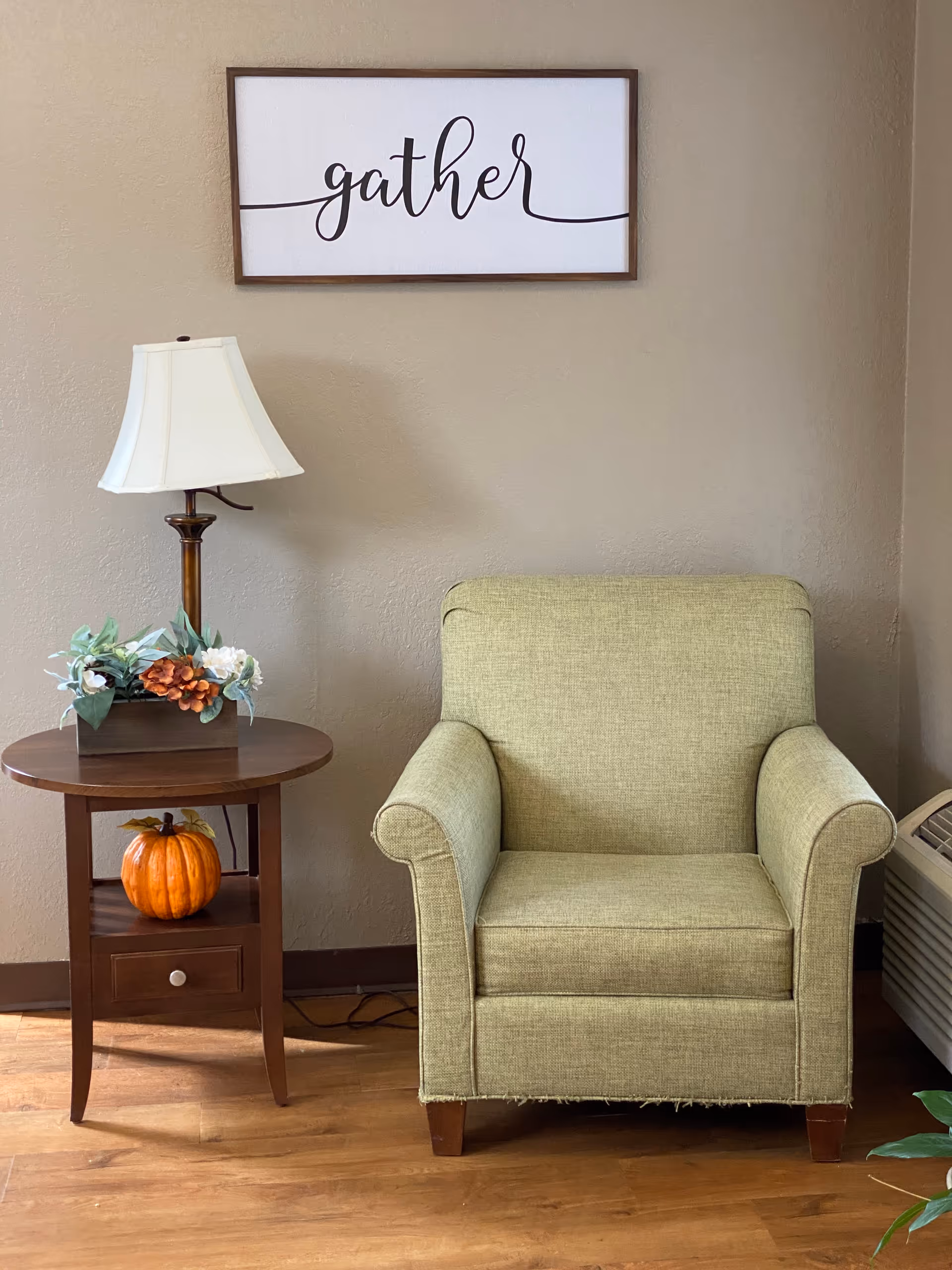 A cozy corner with a light green upholstered armchair next to a wooden side table. The table holds a lamp with a white shade, a floral arrangement, and a small decorative pumpkin on the lower shelf. Above the chair, a framed sign with the word 'gather' is hung on a beige wall. The floor is wooden, and part of an air conditioning unit is visible on the right side.