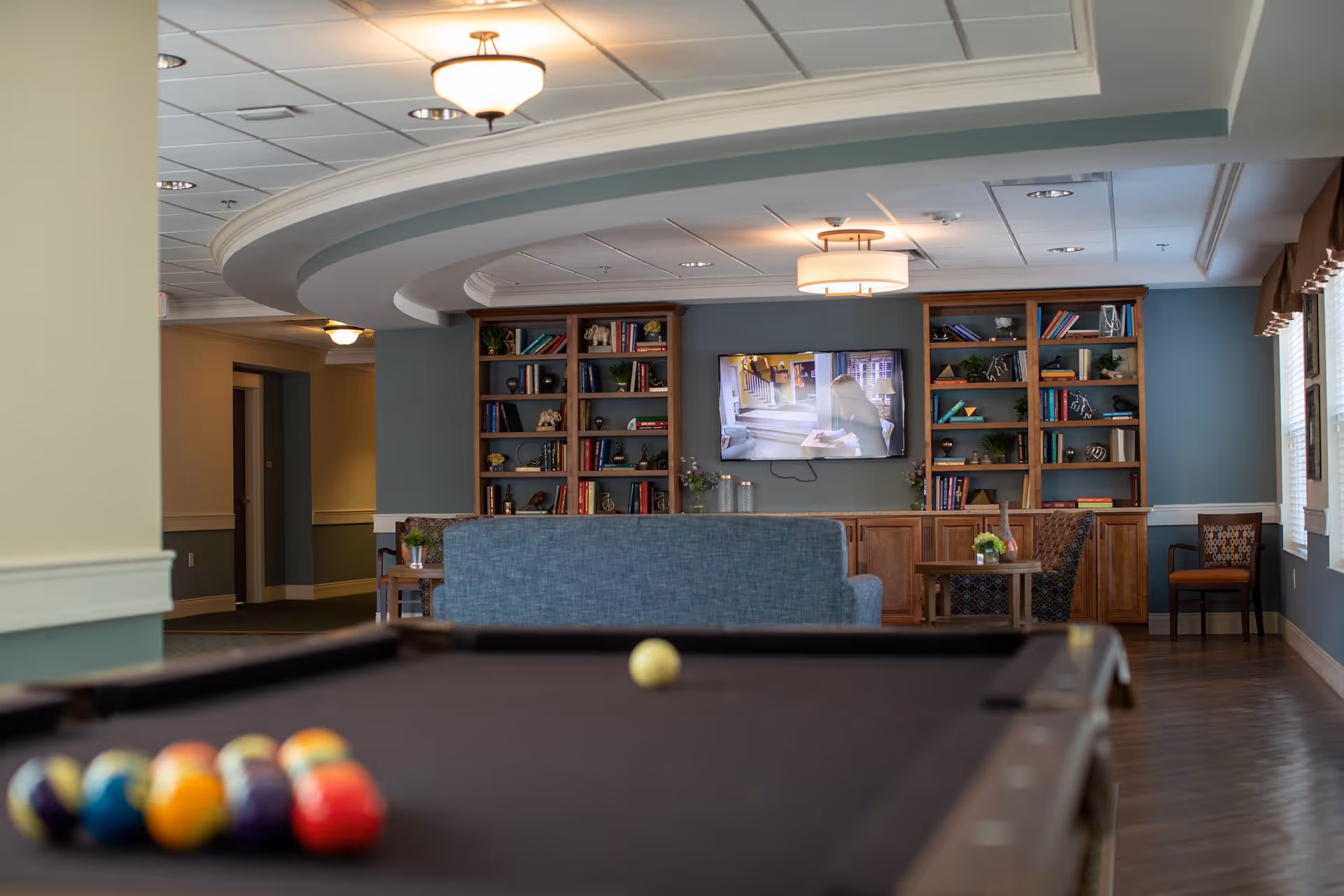 Interior view of a senior living facility common area featuring a pool table in the foreground, a blue couch, wooden bookshelves filled with books and decorative items, a wall-mounted TV, and several chairs near windows with brown valances.