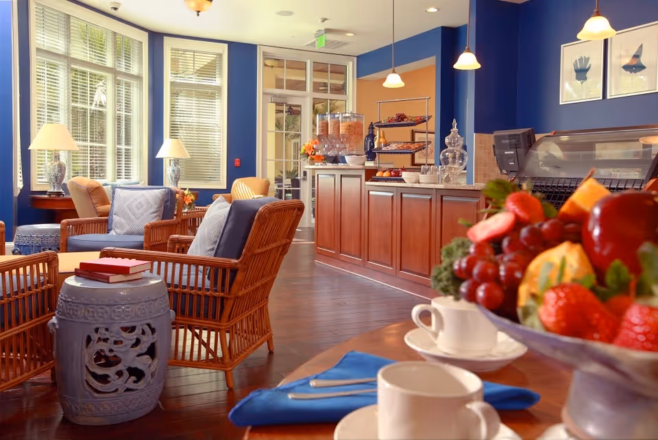 Bright communal dining and lounge area with wicker chairs, a wooden service counter, and a bowl of fresh fruit in the foreground.