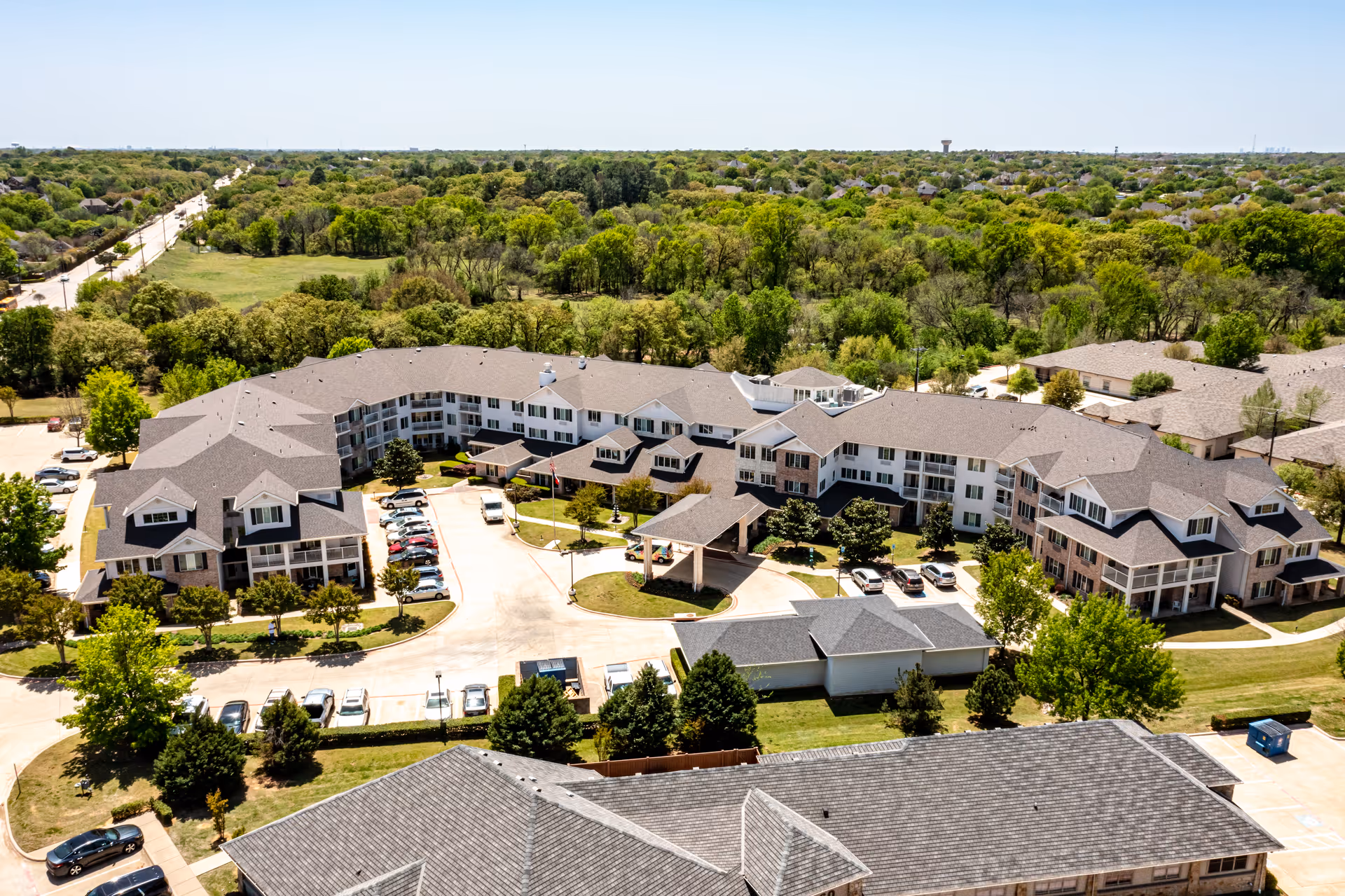 Aerial view of a large L-shaped senior living facility with parking areas, driveways, and surrounding trees and lawns.