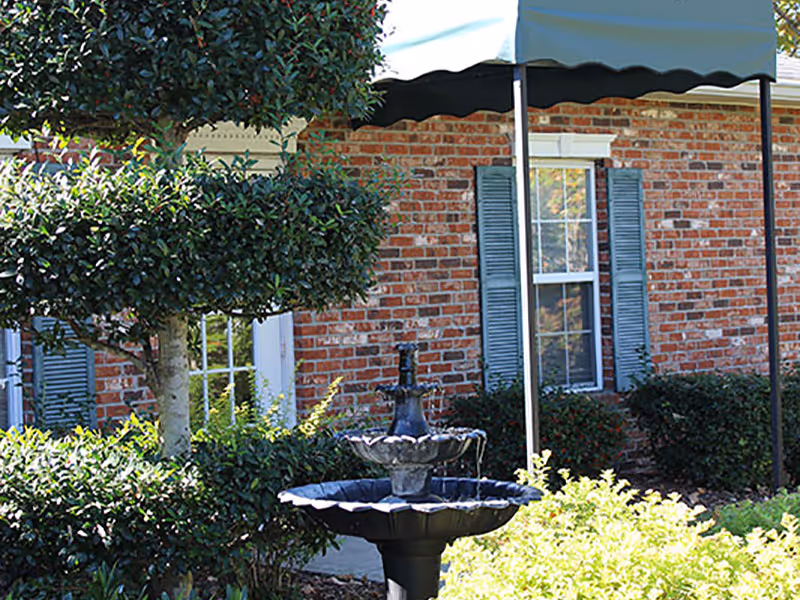 A landscaped courtyard with a tiered fountain in front of a brick building with windows and a green awning.