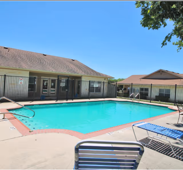 Fenced outdoor swimming pool with lounge chairs in front of single-story residential buildings under a clear blue sky.