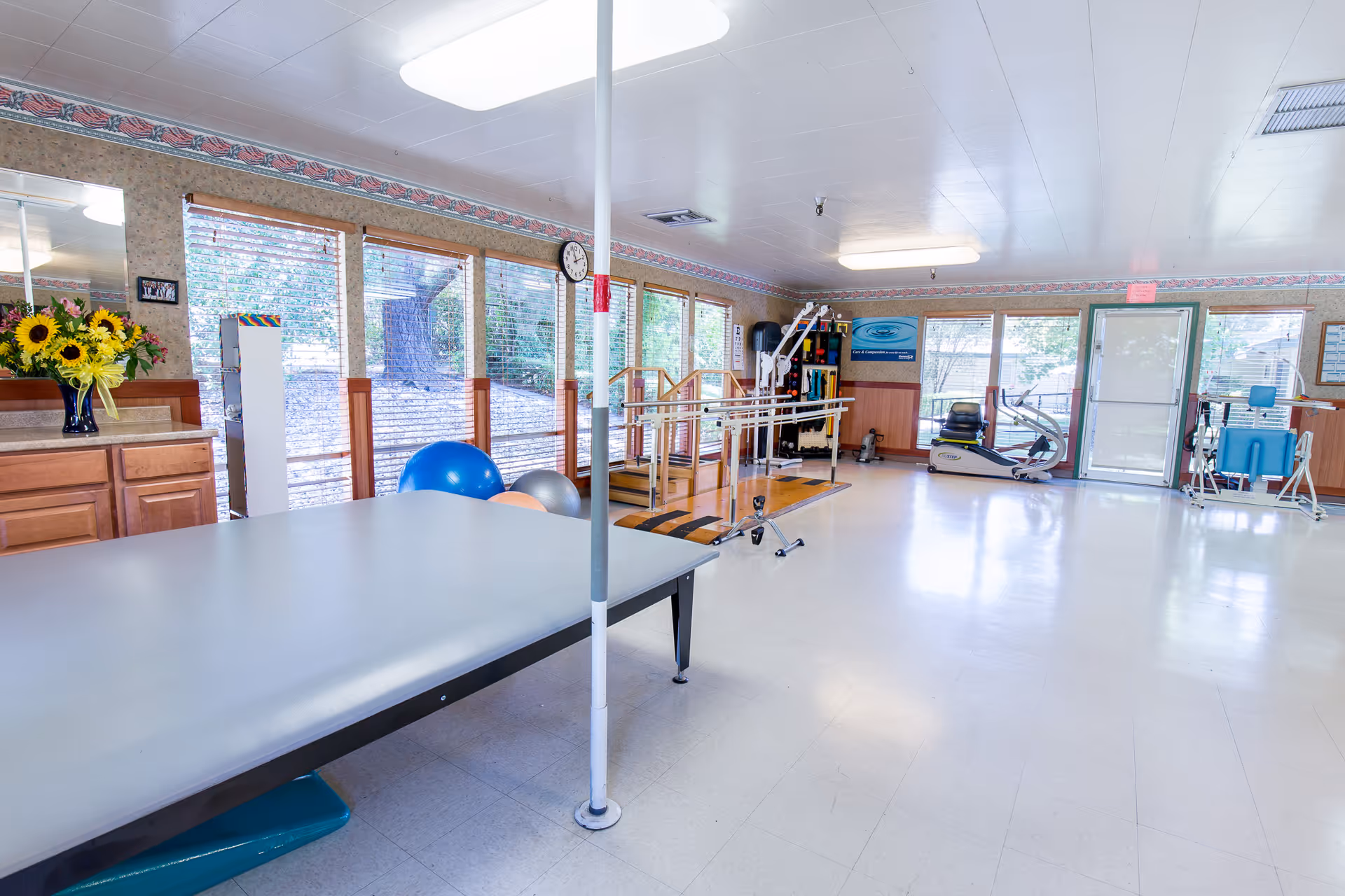 A bright physical therapy or rehabilitation room with large windows along one wall letting in natural light. The room contains exercise equipment including parallel bars, therapy balls, a treatment table, and various fitness machines. There is a vase with sunflowers on a wooden cabinet and a clock on the wall.