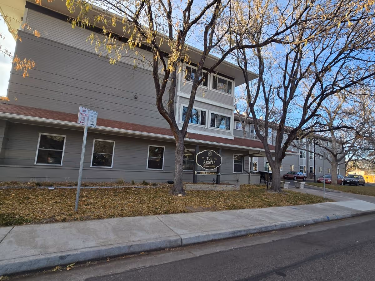 Street-level view of The Argyle building with its entrance sign, leafless trees, sidewalk, and parked cars.