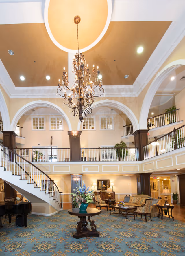 Spacious and elegant senior living facility lobby with a large ornate chandelier hanging from a high ceiling with arches. The room features a curved staircase with white railings, a grand piano, a round table with a floral arrangement, and comfortable seating areas with sofas and armchairs. The decor includes warm lighting, plants, and classic furniture on a patterned blue carpet.
