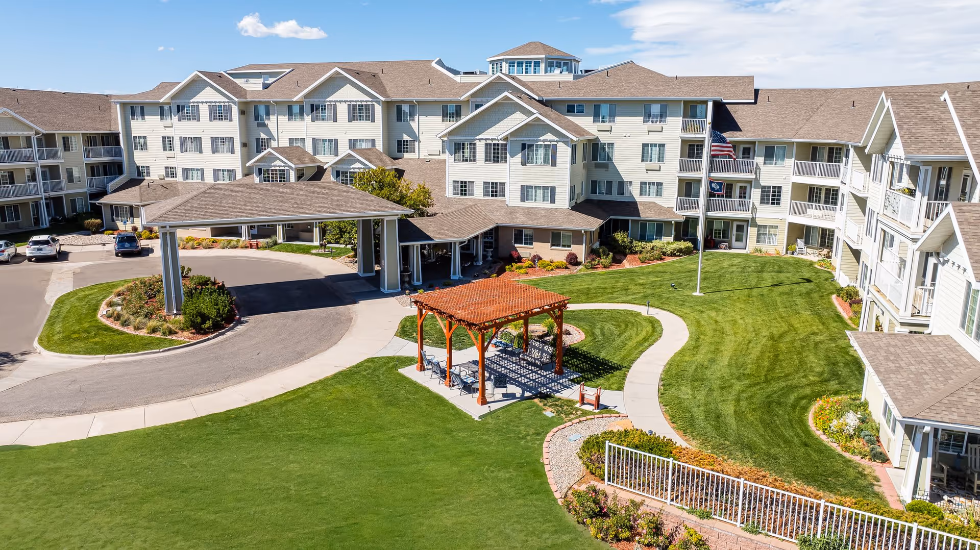 A large senior living facility with multiple stories and beige siding, surrounded by well-maintained green lawns and landscaped gardens. In front of the building is a covered driveway for vehicle drop-off, a paved circular driveway, and a wooden pergola with seating underneath. The sky is clear with a few clouds.