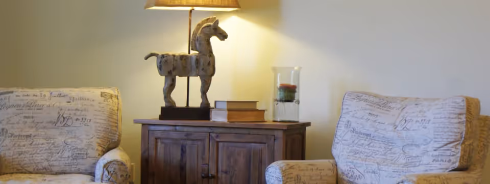 Two patterned armchairs flank a wooden side cabinet topped with a horse lamp, books, and a glass candle holder.