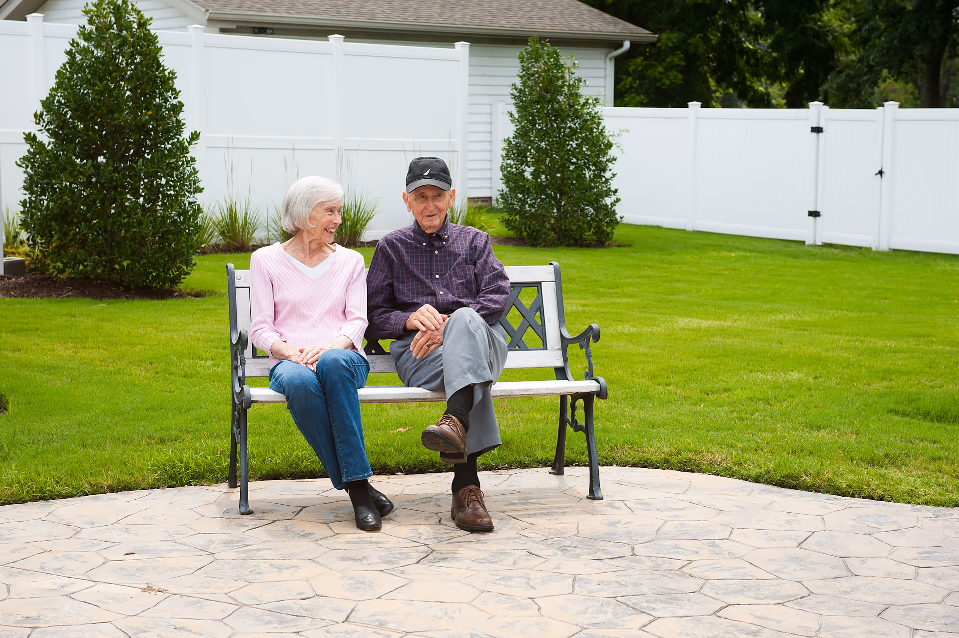 An elderly man and woman sitting on a metal and wood bench in a well-maintained outdoor garden area with green grass, bushes, and a white fence in the background. The woman is wearing a pink sweater and blue jeans, and the man is wearing a dark plaid shirt, gray pants, brown shoes, and a black cap. They appear to be engaged in a pleasant conversation.