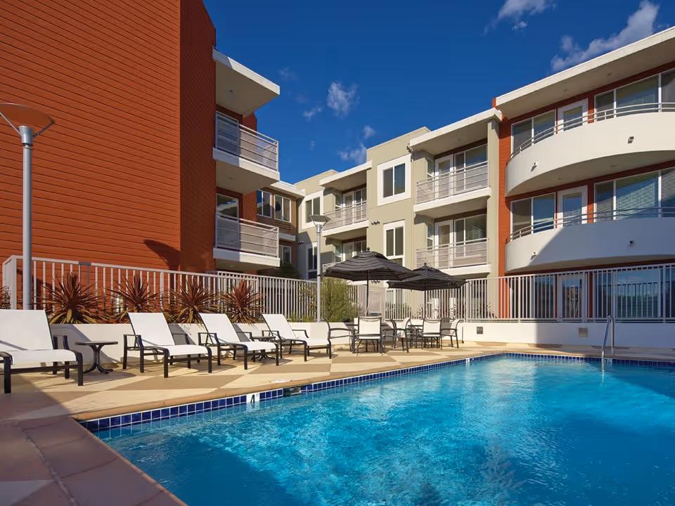 Outdoor swimming pool area at NOHO Senior Arts Colony Apartments with lounge chairs and tables with umbrellas, surrounded by a modern multi-story apartment building under a clear blue sky.