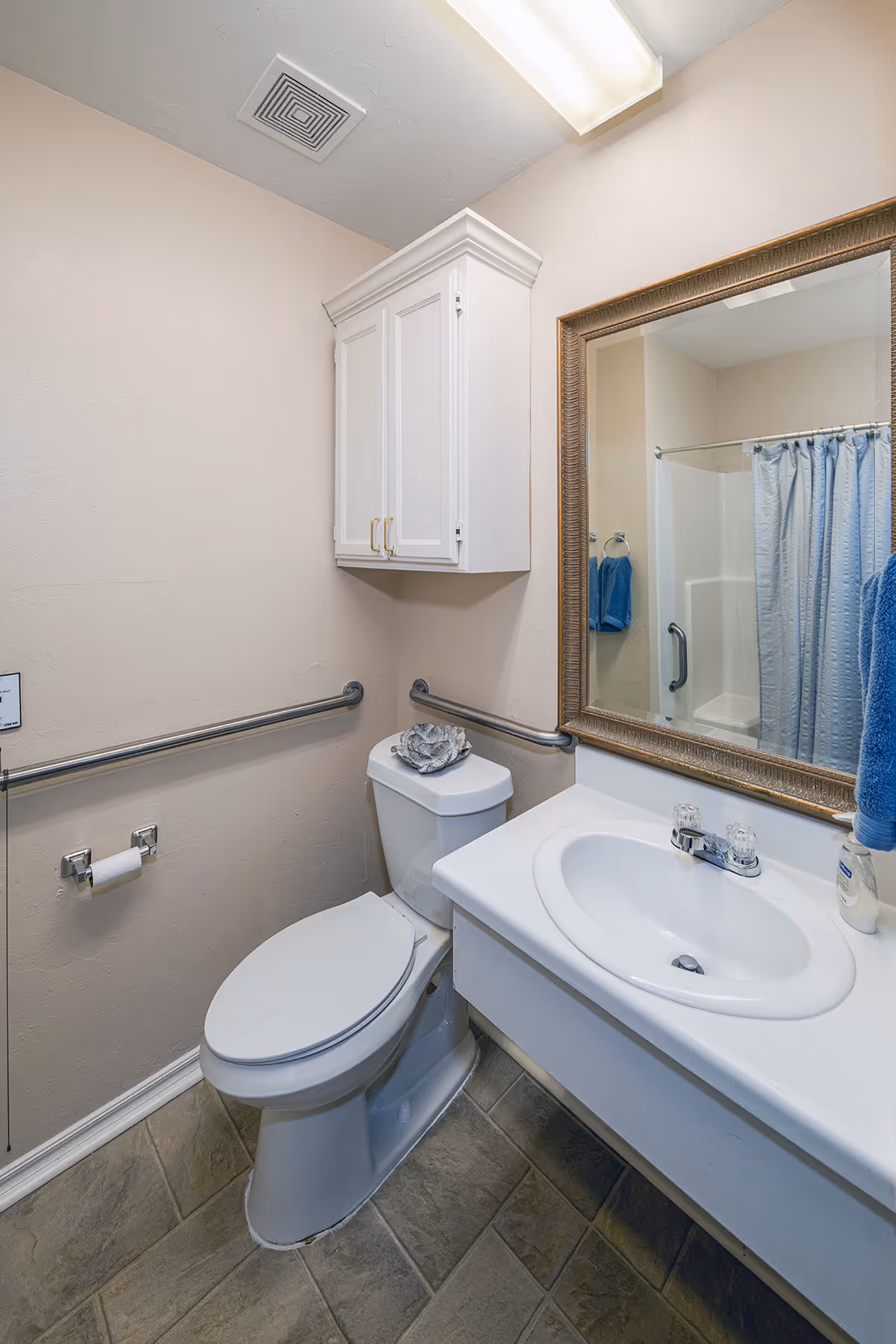 A small bathroom with a white toilet, a white sink with a faucet, a large framed mirror above the sink, and a white cabinet mounted on the wall. There are metal grab bars on the walls near the toilet and sink, a blue towel hanging on a ring, and a shower with a blue curtain visible in the mirror reflection.