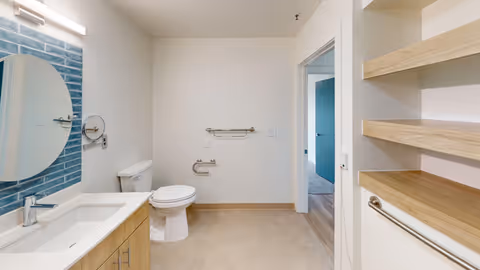 A clean and modern bathroom featuring a white toilet, a wooden vanity with a white sink, a round mirror mounted on a blue tiled wall, and open wooden shelves on the right. The bathroom has beige floor tiles and a doorway leading to another room with wooden flooring.