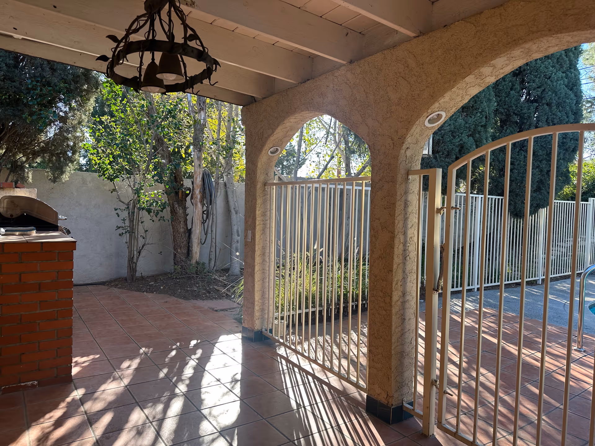 Covered tiled patio with arched stucco columns, a metal security gate, a brick countertop/grill, and trees beyond.